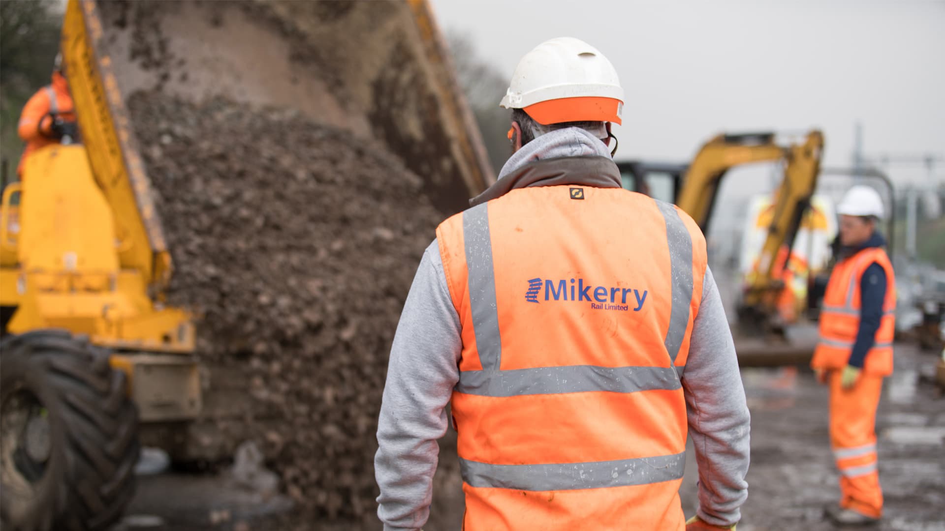 A Mikerry Rail track worker in branded hi-vis watching ballast tip from a dumper, with a second crew member and excavators on the worksite behind